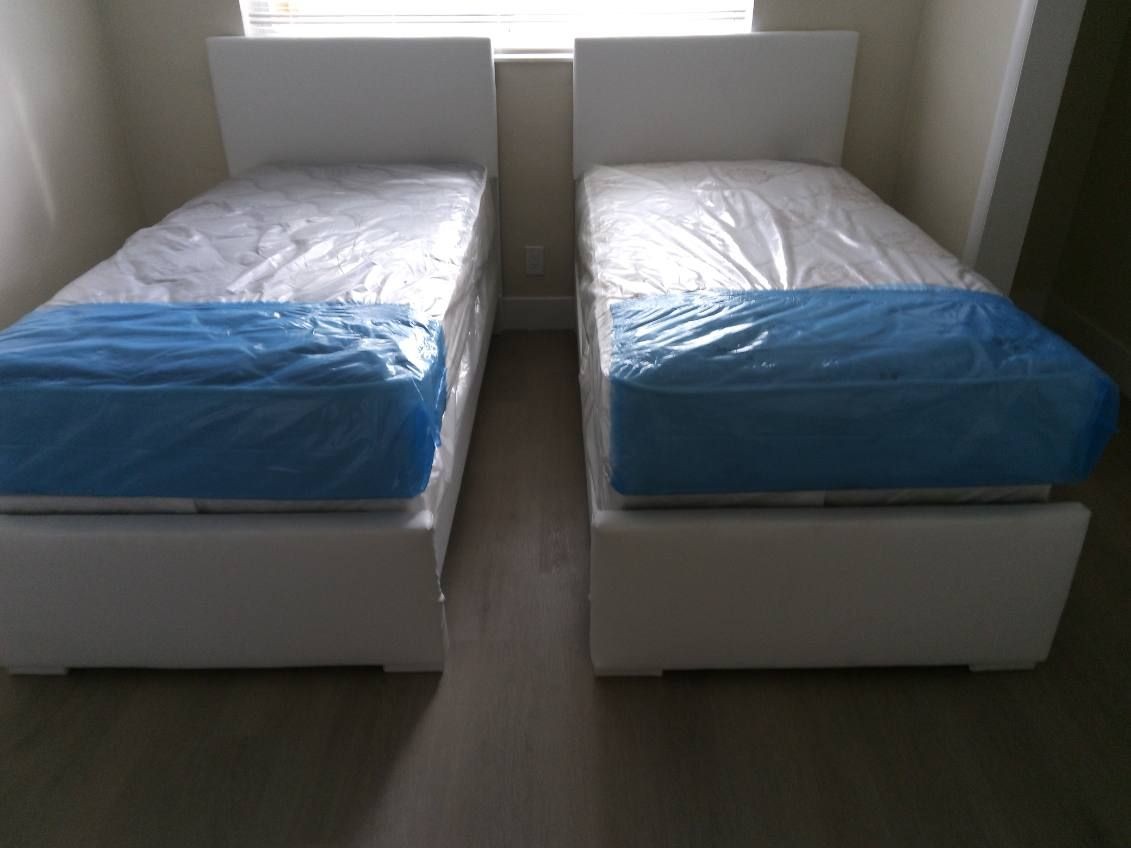 Two twin beds with white mattresses covered in blue protective plastic in a dimly lit room.